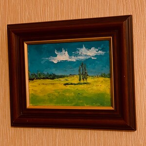 May include: A framed painting of a landscape with a blue sky, white clouds, and a field of green and yellow grass. There are three trees in the distance.