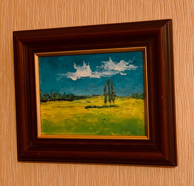 May include: A framed painting of a landscape with a blue sky, white clouds, and a field of green and yellow grass. There are three trees in the distance.
