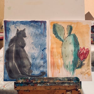 May include: Two watercolor paintings on display. The left painting features a black cat silhouette against a blue background with white splatters. The right painting shows a green cactus with a red flower against a yellow background. The text "echoes" is written on the left painting and "chilla time" is written on the right painting.