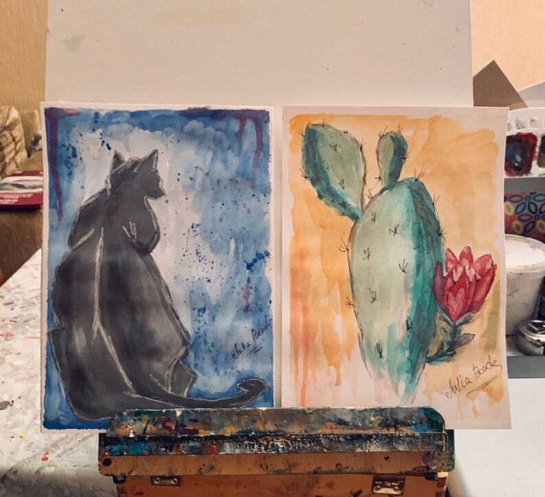 May include: Two watercolor paintings on display. The left painting features a black cat silhouette against a blue background with white splatters. The right painting shows a green cactus with a red flower against a yellow background. The text "echoes" is written on the left painting and "chilla time" is written on the right painting.