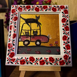 May include: A framed painting of three black cats in a red car with a black cat on top of the car. The background is gold with red flowers. The frame is white with a red and white floral pattern.