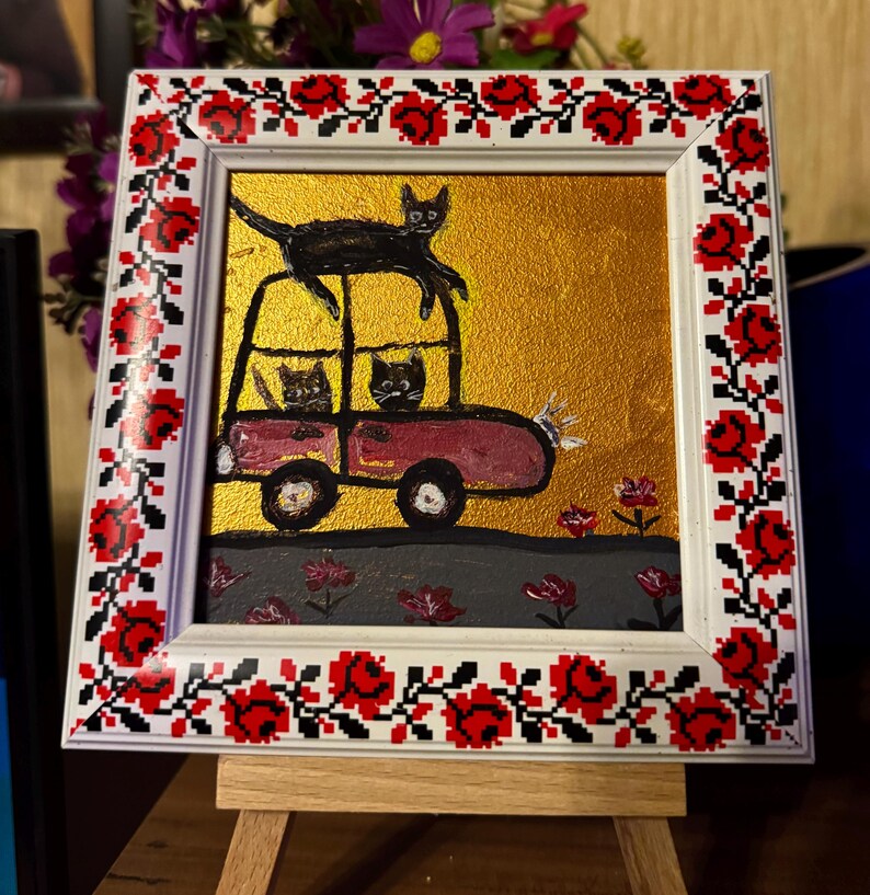 May include: A framed painting of three black cats in a red car with a black cat on top of the car. The background is gold with red flowers. The frame is white with a red and white floral pattern.