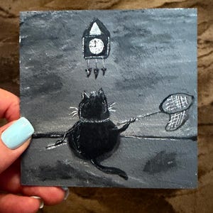 Puede incluir: Una pintura caprichosa de un gato negro sosteniendo una red de mariposas, con un reloj de cuco encima. La obra de arte est&aacute; sobre un fondo cuadrado gris. El gato tiene bigotes blancos y un collar. El reloj tiene una esfera blanca.
