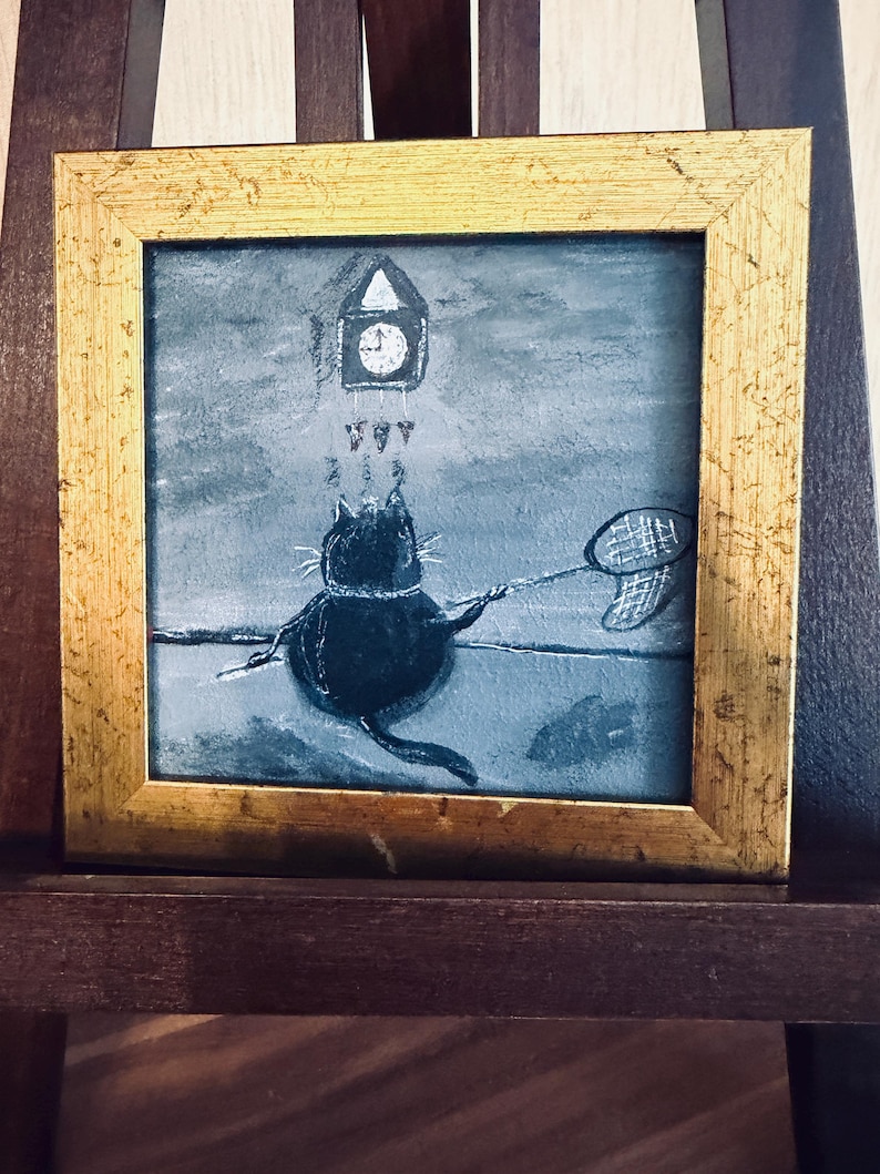 Puede incluir: Pintura enmarcada cuadrada con un marco dorado. La obra de arte representa un gato negro con una red de mariposas, mirando un reloj. El fondo es azul y gris apagado. La pintura se exhibe en un caballete de madera.