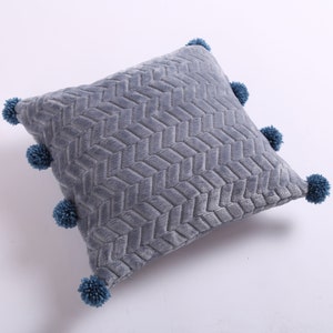 May include: A gray throw pillow with a textured woven pattern and blue pom-poms around the edges.