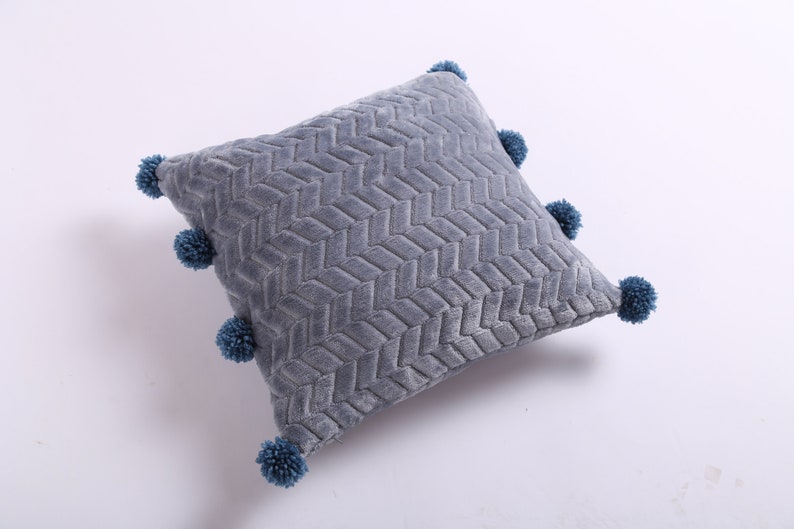 May include: A gray throw pillow with a textured woven pattern and blue pom-poms around the edges.