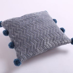 May include: A gray throw pillow with a woven pattern and blue pom-poms around the edges.