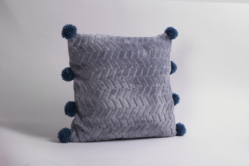 May include: A gray throw pillow with a textured chevron pattern and blue pom-poms around the edges.