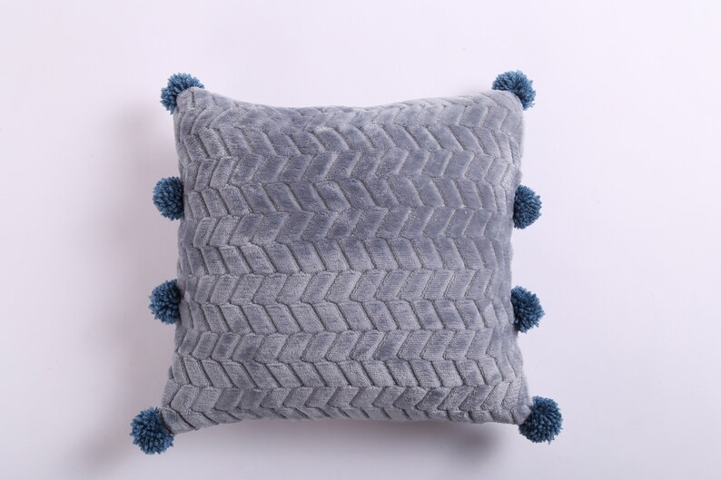 May include: A gray throw pillow with a textured chevron pattern and blue pom-poms around the edges.