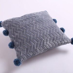 May include: A gray throw pillow with a woven pattern and blue pom-poms around the edges.