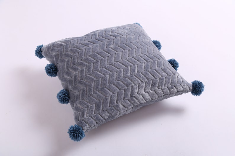 May include: A gray throw pillow with a woven pattern and blue pom-poms around the edges.