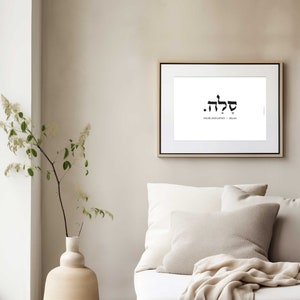 May include: A framed print with the Hebrew word "Shalom" in black lettering on a white background. The word "Shalom" is translated as "peace and justice" in English. The print is hanging on a light beige wall above a white sofa with beige throw cushions.
