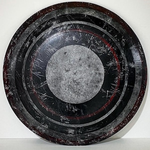 May include: A distressed, circular shield with concentric rings of black, gray, and red. The center is a lighter gray with a scratched texture. The shield has a weathered appearance, suggesting use or age. The overall design is reminiscent of a superhero's shield.