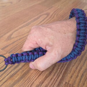 May include: A blue and purple paracord wrist lanyard with a black metal ring. The lanyard is braided and wrapped around a wrist. The background is a wooden surface.