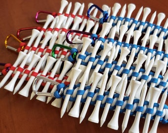 May include: Close-up of golf tee holders made of white tees woven together with red, blue, and white paracord. Each holder has a carabiner clip in various colours. The holders are designed to keep golf tees organised.