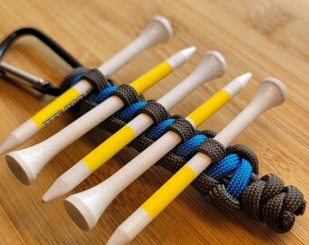 May include: A golf tee holder featuring six white wooden tees with yellow stripes, held together by a woven cord in black, blue, and grey. A black carabiner clip is attached. The text "DTS PRO" is visible on one tee.