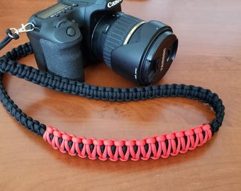 May include: A black camera strap with a red braided section. The strap is made of paracord and is designed to be durable and comfortable.