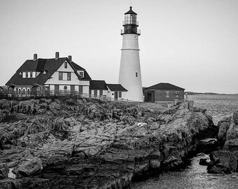 Può includere: Una fotografia in bianco e nero di un faro su una costa rocciosa. Il faro è alto e bianco con un tetto nero. Le rocce sono irregolari e accidentate. L'acqua è calma e il cielo è nuvoloso.