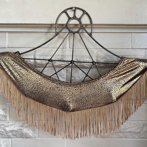May include: A decorative wall hanging featuring a gold and brown patterned fabric with long, beige fringe. The fabric is draped over a black metal frame with a circular top and geometric design, creating a unique, eye-catching piece.