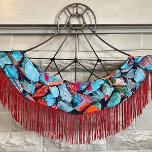 May include: A decorative wall hanging featuring a black metal frame with a curved, fabric-covered section. The fabric displays a pattern of blue, orange, and brown stones. Red fringe hangs from the bottom, adding a textural element.