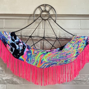 May include: A colorful, abstract print fabric neck scarf with a pink fringe trim. The scarf is hanging on a black metal hanger.
