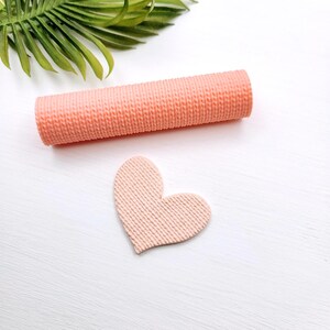 May include: A cylindrical peach-coloured rolling pin with a fine, textured knit-like pattern and a matching heart-shaped cutout piece featuring the same consistent, raised, linear surface texture throughout the entire design.