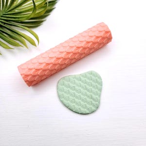 May include: A cylindrical peach-coloured rolling pin featuring a raised, repeating scale texture, accompanied by a small, irregular piece of light green clay that displays the same embossed scale pattern across its entire surface.