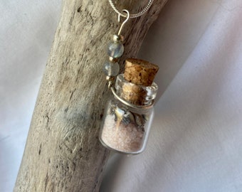 May include: A silver chain necklace with a small glass bottle pendant filled with pink sand and seashells. The bottle has a cork stopper and is attached to the chain with a silver wire. Three light blue beads are on the chain above the bottle.