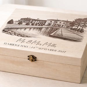 Puede incluir: Una caja de madera de color claro con tapa con bisagras y cierre metálico. La tapa presenta una ilustración grabada detallada de un edificio con un estanque y árboles. También está grabado el texto "Mr & Mrs Mills, Elmridge Hall, 24th September 2027".