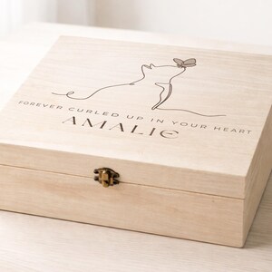 May include: A light-colored wooden box with a hinged lid and a metal clasp. The lid features a line drawing of a cat with a butterfly and the text "FOREVER CURLED UP IN YOUR HEART" and the name "AMALIE".