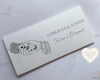 May include: White card with black text that reads "Congratulations Jasleen & Manpreet". A black line drawing of two hands holding each other with a ring on one finger. The card also has a heart shape with the text "Personalised message inside".