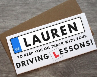 May include: A white card with a blue and white number plate design. The number plate says "GB LAUREN" and the text below reads "To keep you on track with your driving lessons!"  A red "L" learner driver symbol is on the card.  The card has a brown envelope behind it.