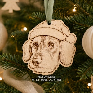 May include: A light-colored wooden ornament shaped like a dog wearing a Santa hat, featuring a detailed laser-engraved portrait of a dog. The ornament hangs from a sage green ribbon and includes the text "PERSONALIZE WITH YOUR OWN PET".