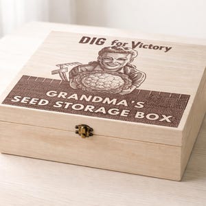 May include: A light-coloured wooden box with a hinged lid and a metal clasp. The box is decorated with a vintage-style graphic that includes the text "DIG for Victory" and "GRANDMA'S SEED STORAGE BOX". The graphic features an illustration of a smiling person holding a large vegetable.