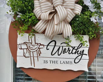May include: A round wooden sign with a rustic white background and a brown border. The sign features a cross with a crown of thorns and the text "Worthy is the Lamb." The sign is decorated with a tan and white striped bow and greenery.
