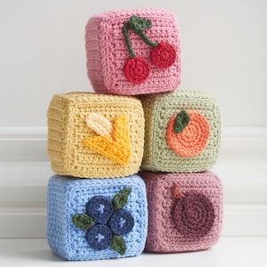 May include: A stack of six crocheted baby blocks in various colors, each adorned with a different fruit. The fruits include cherries, a banana, an orange, blueberries, and a plum. The blocks are stacked on top of each other.