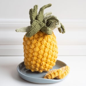 Pode incluir: Um abacaxi de crochê com corpo amarelo e folhas verdes. O abacaxi está sentado em um prato cinza com uma fatia de abacaxi ao lado.
