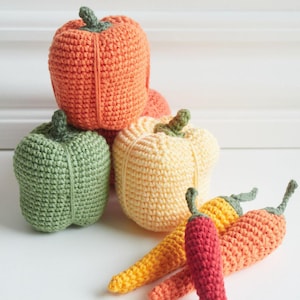 May include: A collection of handmade crochet vegetables, including orange, green, and yellow bell peppers, and red, orange, and yellow chili peppers. The peppers have green stems and are arranged on a white surface.