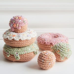 Puede incluir: Una colección de donuts y una magdalena de ganchillo hechos a mano en varios tamaños y colores. Los donuts tienen una base marrón claro con glaseado blanco, rosa y verde, y chispas de colores. La magdalena es rosa con chispas.
