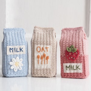 May include: Three handmade crochet milk carton covers. One is light blue with a daisy and the word "MILK". The second is beige with the word "OAT" and embroidered wheat. The third is pink with a strawberry and the word "MILK".