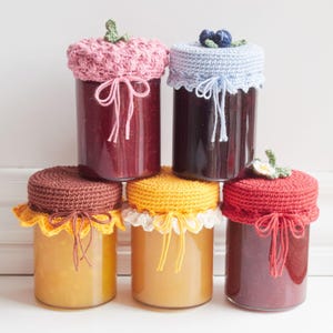 May include: Six glass jars filled with preserves, each topped with a unique crocheted lid. The lids are in various colors, including pink, blue, brown, yellow, and red, and are tied with matching yarn. Some lids have decorative embellishments.