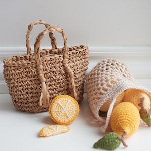 Puede incluir: Un bolso tote de paja tejida con asas trenzadas y borlas se encuentra junto a una bolsa de ganchillo llena de limones de ganchillo amarillos. También hay dos rodajas de limón de ganchillo. Los artículos están dispuestos sobre una superficie blanca.