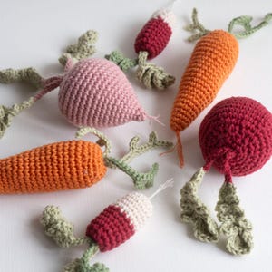 Puede incluir: Un conjunto de cinco verduras de ganchillo, que incluyen una remolacha rosa, dos zanahorias naranjas y dos rábanos rojos. Las verduras están todas hechas a mano y tienen hojas verdes de ganchillo.