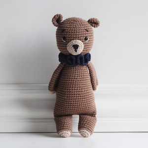 Puede incluir: Un oso de peluche de crochet marrón con un cuello de volantes azul. El oso tiene ojos negros y una nariz negra.