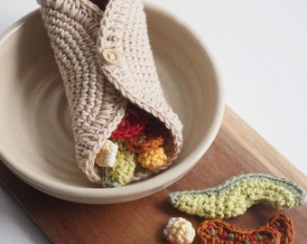 Könnte beinhalten: Ein gehäkelter Burrito mit verschiedenen farbigen Füllungen, darunter Rot, Orange, Gelb und Grün, liegt in einer beigefarbenen Schüssel. Der Burrito ist aus beigefarbenem Garn gefertigt und hat einen Knopfverschluss. Die Schüssel steht auf einer Holzoberfläche mit anderen gehäkelten Lebensmitteln, darunter eine grüne Avocado, eine rote Tomate und ein brauner und orangefarbener Taco.