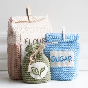 Puede incluir: Tres bolsas de crochet, una beige con la palabra "FLOUR" bordada, una azul con la palabra "SUGAR" bordada y una verde con dos hojas bordadas.