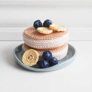 Puede incluir: Una pila de panqueques de crochet con rodajas de plátano y arándanos en la parte superior, exhibida en un plato azul claro.