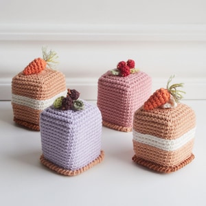 Puede incluir: Cuatro mini tartas de crochet con diferentes coberturas. Las tartas son cuadradas y tienen una capa de glaseado blanco. Las coberturas incluyen una zanahoria, bayas y un grupo de pequeñas bayas moradas y marrones.