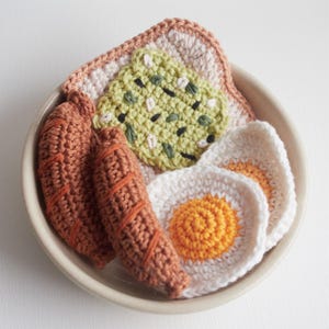 Peut inclure: Un ensemble de petit-déjeuner crocheté dans un bol blanc. L'ensemble comprend deux saucisses brunes, deux œufs blancs avec des jaunes d'orange et un toast à l'avocat vert.