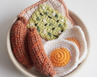 Könnte beinhalten: Ein gehäkeltes Frühstücksset in einer weißen Schüssel. Das Set enthält zwei braune Wurststücke, zwei weiße Eier mit orangefarbenen Dottern und einen grünen Avocado-Toast.
